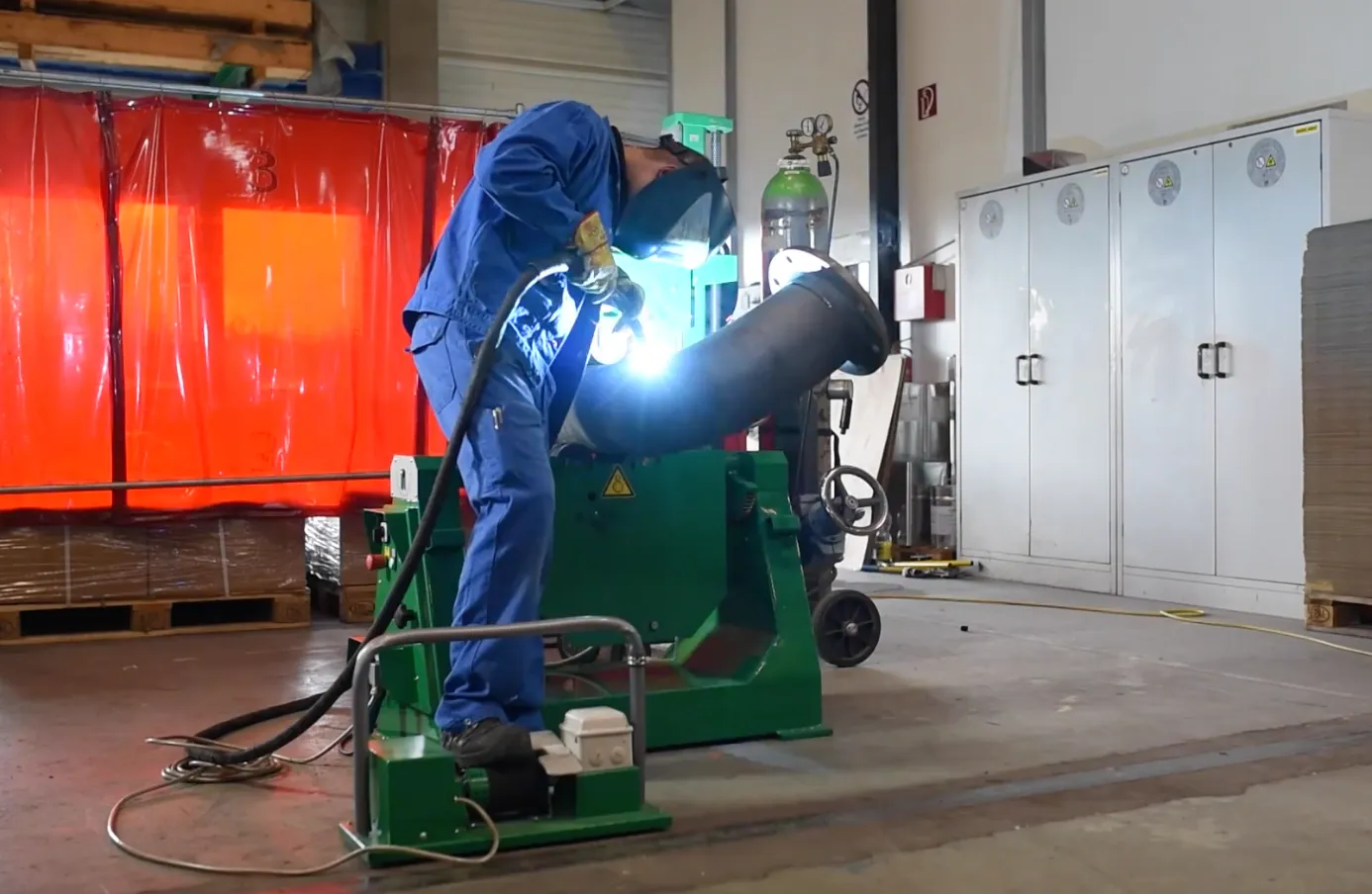 Welder running a continuous pass on a large pipe clamped in a green Kistler U-range pipe rotator in a fabrication shop